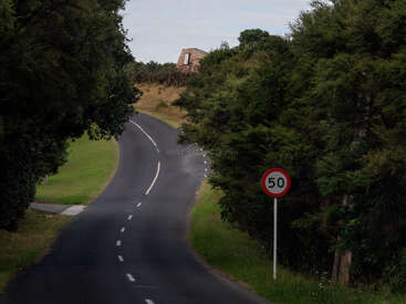 Eine kurvenreiche, asphaltierte Landstraße schlängelt sich durch dichten, grünen Baumbestand. Rechts taucht ein 50-km/h-Tempo-Schild auf. In der Ferne steht ein altes Steinhaus.