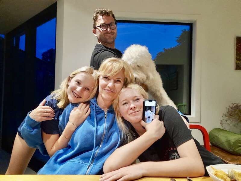 Eine fröhliche Familie versammelt sich um einen Tisch, umarmt sich und lächelt. Im Hintergrund sitzt ein Hund. Ein Mädchen hält ein Handy in der Hand, auf dem ein ähnliches Bild zu sehen ist.