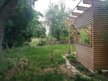 Un jardin paisible avec une végétation luxuriante, des fleurs sauvages, une balançoire en corde suspendue à un arbre et une cabane en bois moderne sur la droite. Clôture en bois en arrière-plan.