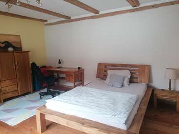 This cozy bedroom features wooden furniture, a double bed, nightstand with lamp, desk with chair, wardrobe, soft rug, exposed ceiling beams, and minimal decor.