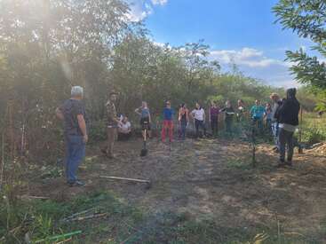 La imagen muestra a un grupo de personas reunidas en una zona boscosa, rodeadas de árboles y arbustos, con algunos individuos sosteniendo herramientas de jardinería.