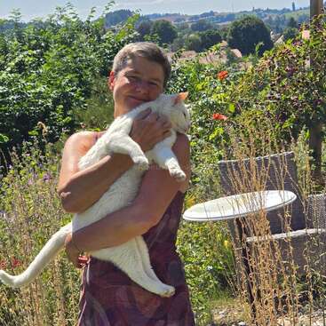 Una mujer de pelo corto abraza cariñosamente a un gran gato blanco en un exuberante jardín iluminado por el sol, lleno de vegetación, flores y muebles de exterior al fondo.