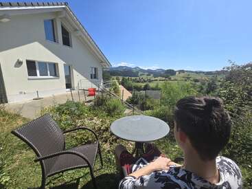 Una persona se sienta al aire libre en una mesa redonda de metal, disfrutando de una vista panorámica del campo junto a una casa moderna, bajo un cielo azul brillante. Ambiente tranquilo.