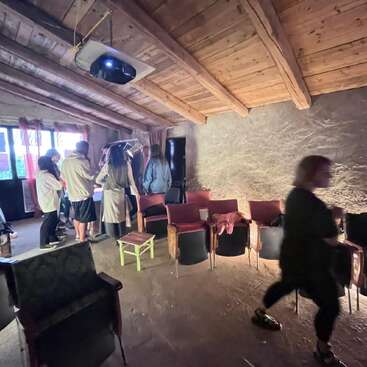 Eine Gruppe von Menschen versammelt sich um einen Tisch in einem rustikalen Raum mit Holzdecken, Stühlen und einem Projektor. Eine Person geht zügig im Vordergrund.