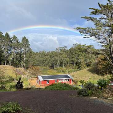 La imagen representa un paisaje sereno con una casa roja con paneles solares, enclavada entre árboles y un vibrante arco iris que se extiende por el cielo.