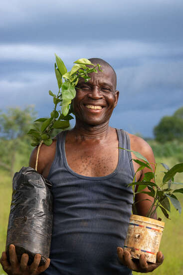Un hombre sonriente está de pie al aire libre, con dos plantones en macetas en las manos. Lleva una camiseta sin mangas oscura y se le nota el orgullo y la alegría que le producen sus labores de jardinería.