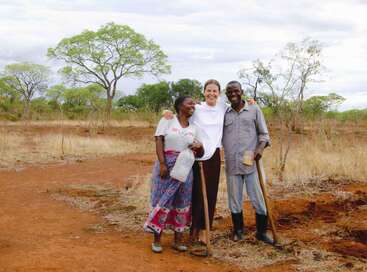 Tres personas posan juntas, sonrientes, en un paisaje rural y árido. Parecen felices, con herramientas agrícolas y un recipiente de agua en las manos, lo que sugiere un trabajo en equipo en tareas agrícolas o comunitarias.