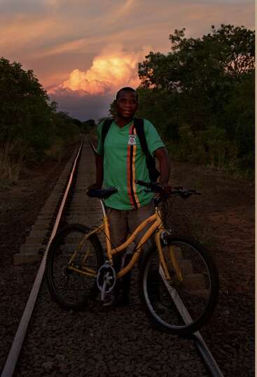 Un hombre está de pie sobre las vías del tren, sosteniendo una bicicleta amarilla. Lleva una camiseta verde y una mochila. El fondo está cubierto de frondosos árboles y espectaculares nubes al atardecer.