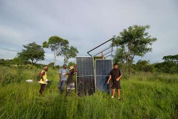 Cinco personas se encuentran de pie entre la hierba alta y verde, montando o revisando dos grandes paneles solares. Al fondo se ven árboles y un cielo azul con nubes, lo que sugiere que se trata de un entorno rural.