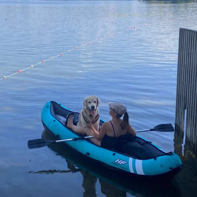 A woman and her dog sit together in a blue kayak on a calm lake, surrounded by trees and blue sky, enjoying a peaceful outdoor day.