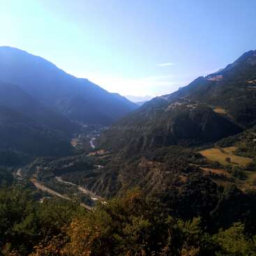 Rolling green hills and valleys stretch beneath a clear blue sky. A winding river snakes through the lush landscape, nestled between majestic mountains and scattered fields.
