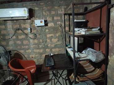 Un petit espace de travail faiblement éclairé avec une chaise en plastique, une étagère en métal, une table avec un clavier, un climatiseur, un ventilateur mural, un mur en briques apparentes et des appareils électroniques et des papiers éparpillés.