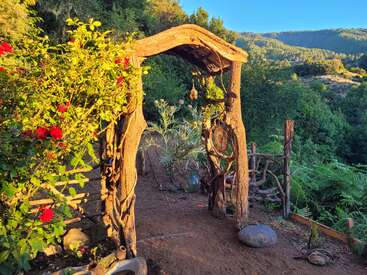 A imagem mostra um arco de madeira rústico em um jardim, adornado com um sino e cercado por uma vegetação exuberante e flores vermelhas vibrantes, em um pitoresco cenário montanhoso.