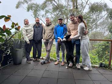 Six people and a black-and-white dog stand together on a patio, surrounded by lush greenery and trees, all smiling and casually dressed outdoors.