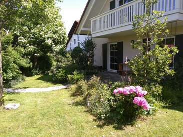 Ein charmantes Haus mit Balkon blickt auf einen üppigen grünen Garten. Im Vordergrund blühen leuchtend rosafarbene Blumen, umgeben von Bäumen und einem friedlichen Weg im Freien.