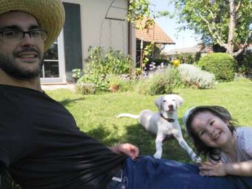 Ein Mann mit einem Strohhut, ein lächelndes Kind und ein weißer Hund entspannen sich gemeinsam auf einem sonnigen, grünen Rasen in einem schönen Garten.