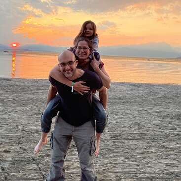 Una familia sonriente disfruta de un momento lúdico en la playa al atardecer. Una persona da paseos a caballito; el cielo resplandece con tonos naranjas y rosas.