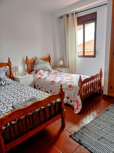 Esse quarto aconchegante tem duas camas de solteiro de madeira com roupas de cama coloridas, um tapete listrado, mesas de cabeceira com luminárias, paredes brancas e uma janela com cortinas creme.