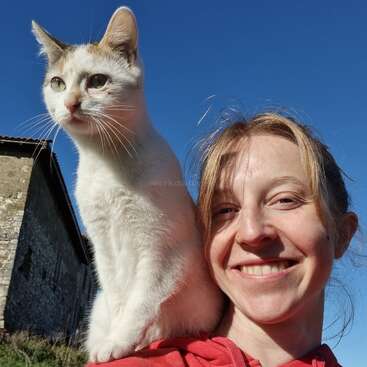 Une femme souriante vêtue d'un sweat à capuche rouge prend un selfie en plein air avec un chat blanc perché sur son épaule, tous deux profitant d'une journée ensoleillée.