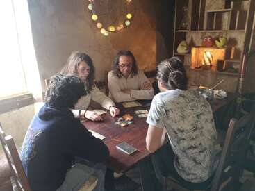 Four people sit around a wooden table playing a board game. The cozy room has warm lighting, decorated shelves, and a string of yellow lights on the wall.