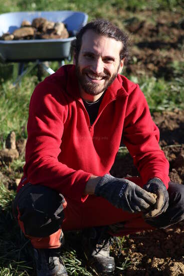 A man with a beard, wearing a red jacket and gloves, kneels outdoors on soil, smiling. Behind him, a wheelbarrow filled with rocks is visible.