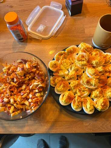 This image shows a wooden table with a bowl of chopped apple and carrot salad, a plate of deviled eggs, a mug, and vitamin bottle.