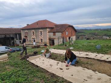 Three people are working together in a garden, planting seeds in rows of prepared earth outside a rustic farmhouse, surrounded by green fields and distant hills.
