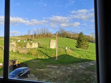 The photo, taken from a window, shows green hills, stone ruins, a black car, a person working, blue sky, and scattered clouds. Peaceful countryside.