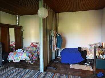 This image shows a shared bedroom with two single beds separated by a curtain, wooden ceiling, wardrobe, clothes rack, rugs, and a small shelving unit.