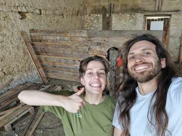 Two people smiling happily in a rustic workshop. One gives a thumbs up. Wooden planks and tools are scattered around, suggesting ongoing DIY or construction work.
