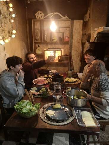 Four people gather around a cozy dinner table. They smile, share food, and take a selfie, under a warm light in a rustic, decorated room.