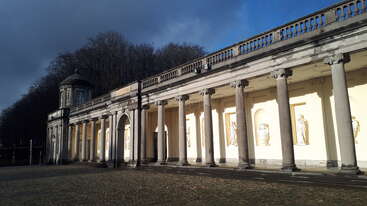 Das Bild zeigt eine klassische Kolonnade mit hohen Steinsäulen, Statuen in Nischen und einem gepflasterten Hof, unter einem dramatischen Himmel und umgeben von dichten Bäumen.