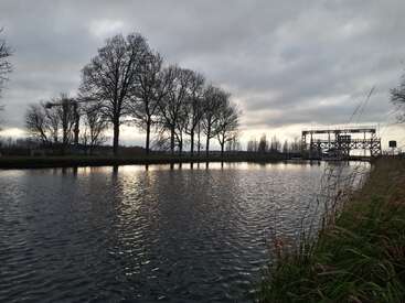 Ein ruhiger Fluss fließt unter einem wolkenverhangenen Himmel und spiegelt die Silhouetten von blattlosen Bäumen und einer Metallbrücke wider. Hohes Gras säumt das Ufer und sorgt für Ruhe.