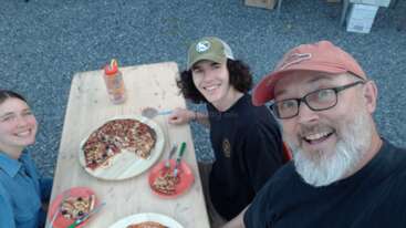 Drei Personen sitzen fröhlich um einen Holztisch im Freien, genießen eine Pizza und lächeln in die Kamera. Auf dem Tisch stehen Teller, Utensilien und eine Wasserflasche.