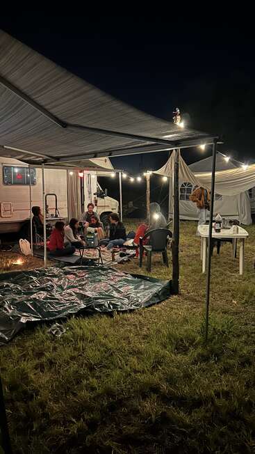 A group of people sits under a canopy, illuminated by string lights. An RV and tents are in the background, creating a cozy nighttime camping scene.
