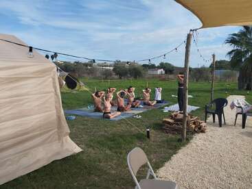 Un grupo de personas practica yoga o meditación al aire libre en colchonetas, siguiendo a un instructor. El entorno es de hierba, con luces de cuerda, sillas y una tienda de campaña.