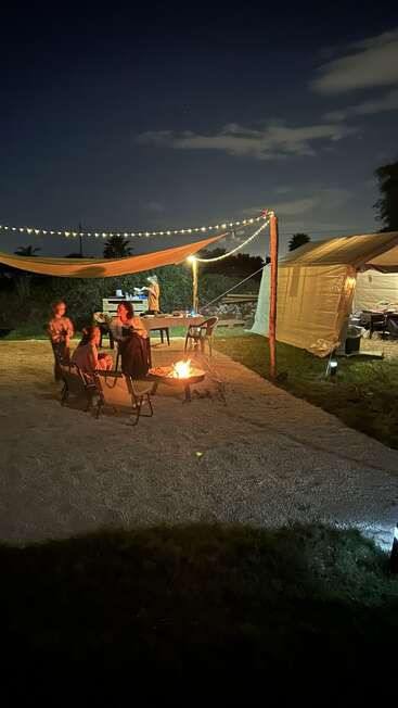 Por la noche, la gente se reúne en torno a una hoguera bajo luces de cuerda y un toldo, con una carpa y una cocina al aire libre visibles al fondo. Ambiente acogedor.