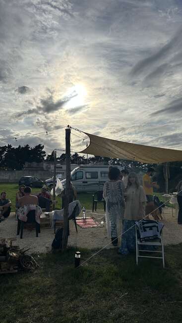 Un grupo de personas se reúne al aire libre bajo un toldo, socializando cerca de una autocaravana. El sol brilla a través de espectaculares nubes, creando un ambiente relajado y comunitario.