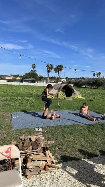 Tres personas practican acroyoga al aire libre sobre una esterilla, con una persona equilibrando a otra sobre sus pies, mientras la tercera se sienta cerca. De fondo, una tienda de campaña y palmeras.