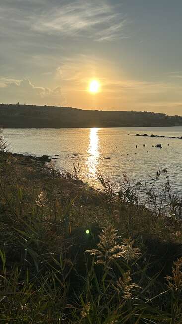 El sol dorado se pone sobre un tranquilo lago, iluminando las suaves olas. Las siluetas de las colinas y la hierba alta en primer plano crean un paisaje natural tranquilo y sereno.