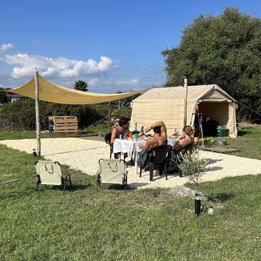 Four people sit around a table eating and talking outside a large beige tent. Two empty chairs face the scene. It’s sunny, relaxed, and outdoorsy.