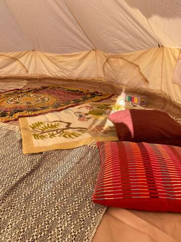 Inside a cozy tent, colorful rugs and pillows create a warm atmosphere. Sunlight filters through, highlighting patterns and textures. A small box sits in the corner.