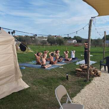 A group of people practice yoga or meditation outside on mats, following an instructor. The setting is grassy, with string lights, chairs, and a tent.