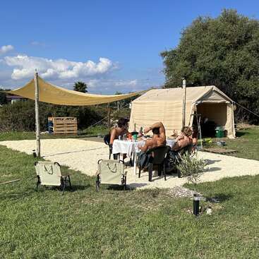 Vier Personen sitzen um einen Tisch herum, essen und unterhalten sich vor einem großen beigen Zelt. Zwei leere Stühle stehen vor der Szene. Es ist sonnig, entspannt, und man ist im Freien.