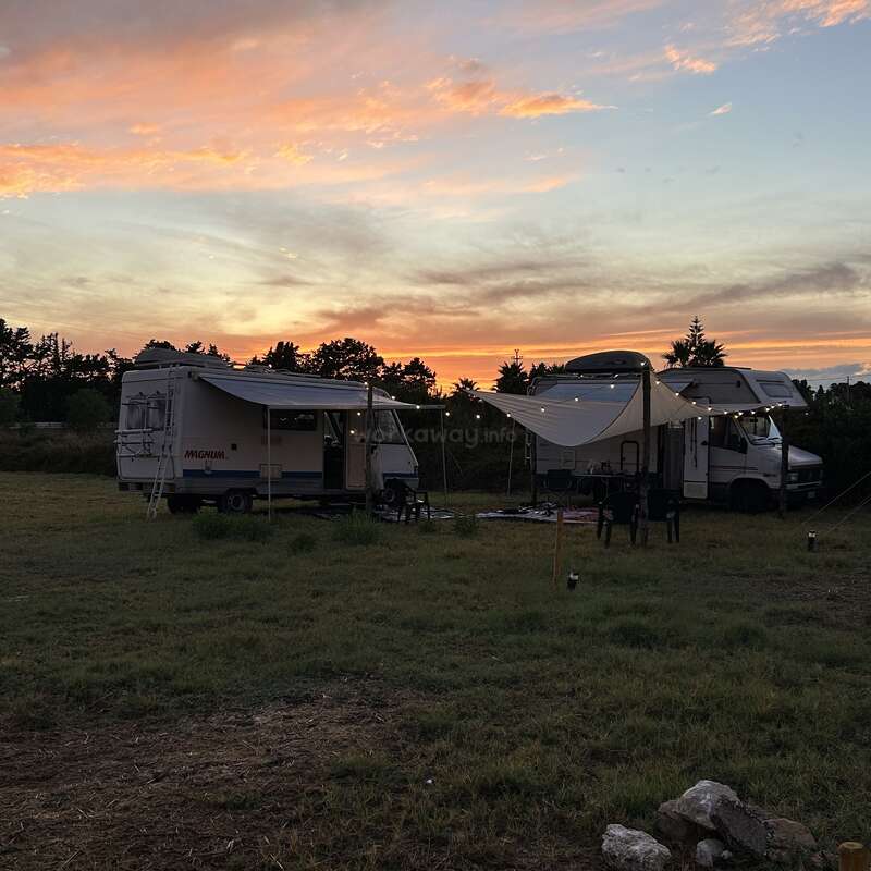 Two camper vans are parked on grassy land at sunset. Warm lights hang under a canopy. Trees silhouette the horizon, creating a peaceful, outdoor camping scene.