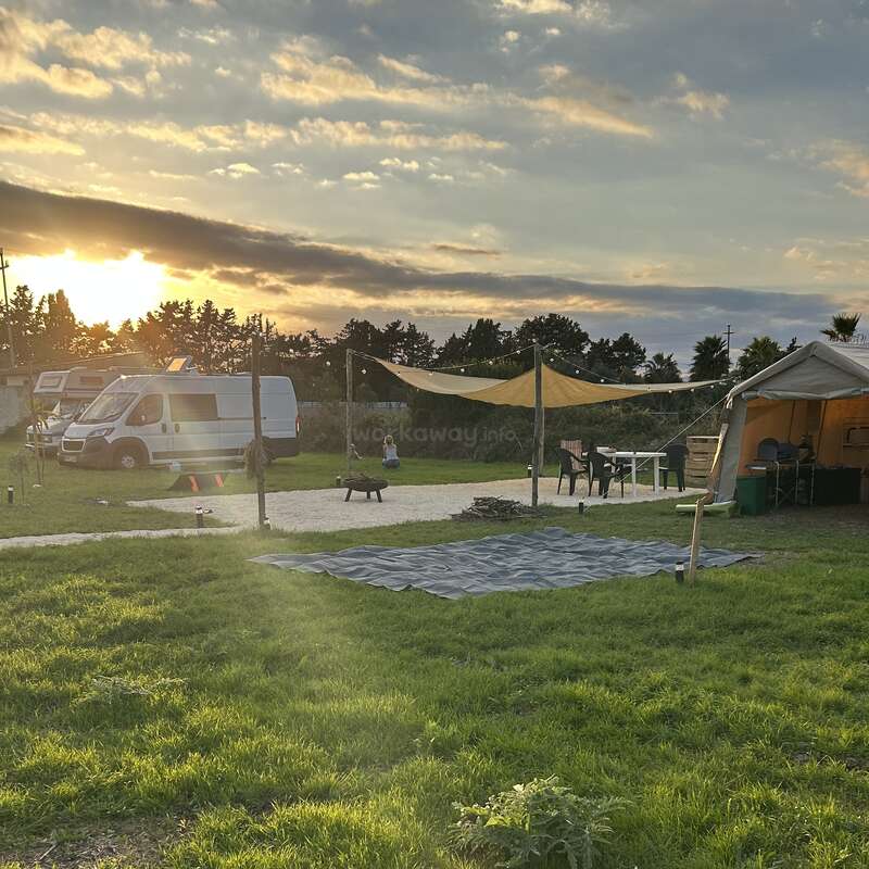 Un tranquilo campamento al atardecer, con una furgoneta, tiendas de campaña, asientos al aire libre, mesas, zona de sombra y exuberante hierba verde. El cielo tiene hermosas nubes y la luz del sol.