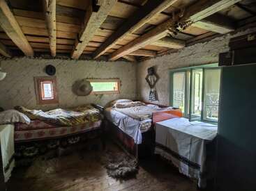 Esta acogedora habitación rústica cuenta con dos camas individuales con mantas de colores, vigas de madera en el techo, un armario verde, adornos tejidos, pequeñas ventanas y muebles sencillos hechos a mano.