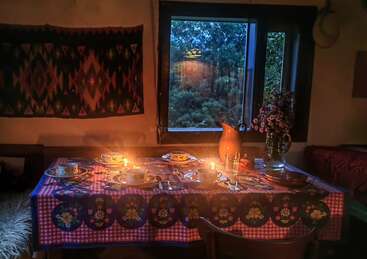 Una acogedora mesa de comedor para cuatro personas, cálidamente iluminada, adornada con un colorido mantel, tazas de té, velas, flores y una gran ventana que deja ver la exuberante vegetación del exterior.