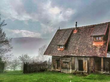 Una rústica casa de madera se asienta sobre la hierba verde, rodeada por una valla de madera. La niebla y los árboles sin hojas crean una atmósfera misteriosa y apacible bajo un cielo nublado.