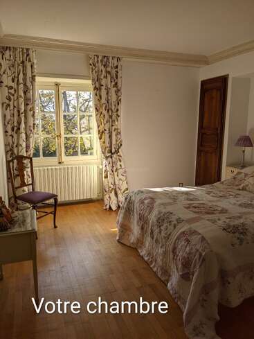 Este acogedor dormitorio cuenta con una cama grande con una colcha de flores, cortinas a juego, suelos de madera, una silla, una ventana, un pequeño escritorio, una lámpara y el letrero «Votre chambre».